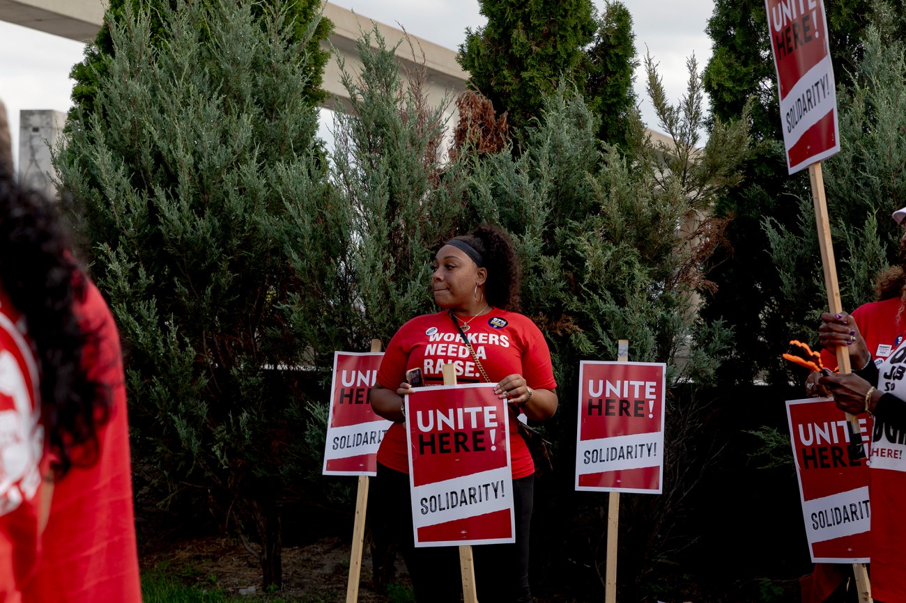 Woman in a red t-shirt standing in front of bushes holding a UAW strike sign.