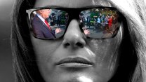 A close-up black-and-white portrait of Melania Trump. Reflected on the lenses of her sunglasses is Donald Trump, who is speaking, with arms outstretched, to a gathering of reporters.