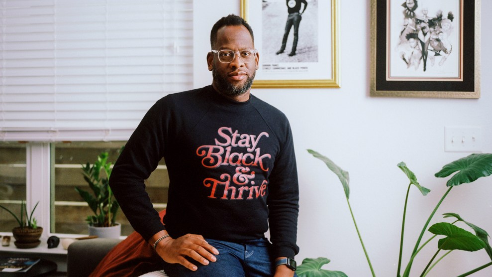 In this photo, Evan Narcisse, a slender Black man is wearing translucent glasses, blue jeans and a black long-sleeve shirt that says "Stay Black and Thrive." On the wall behind him is a framed photo of Stokely Carmichael in a Black Panther Party sweater.