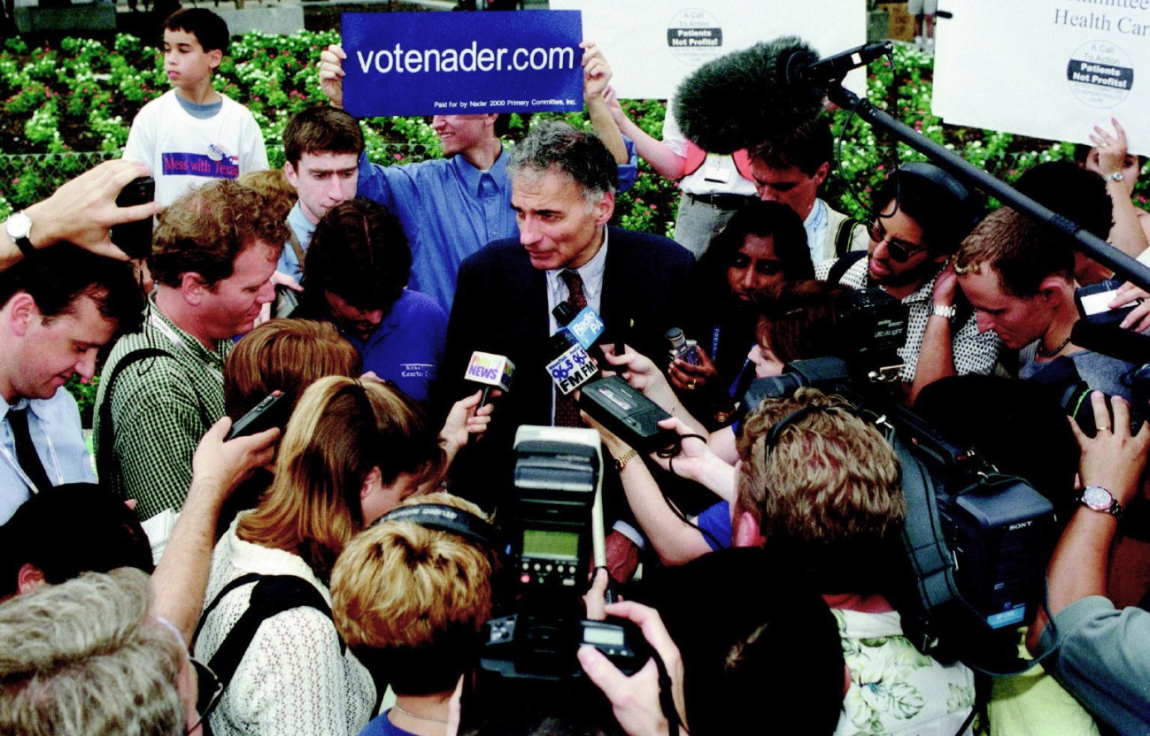 Ralph Nader surrounded by reporters with microphones and cameras.