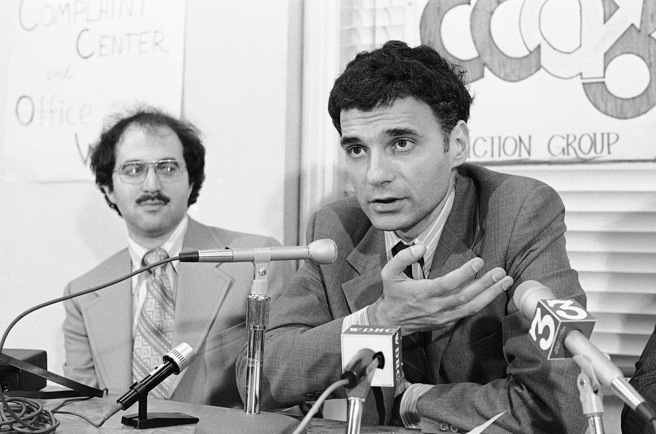 Black and white photo of Ralph Nader speaking at microphones, seating next to a man with a mustache and glasses.