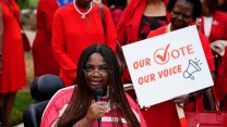A Black woman wearing red in a wheelchair speaking into a microphone wearing red, surrounded by other Black woman wearing red, including one carrying a poster that says "Our Vote, Our Voice"