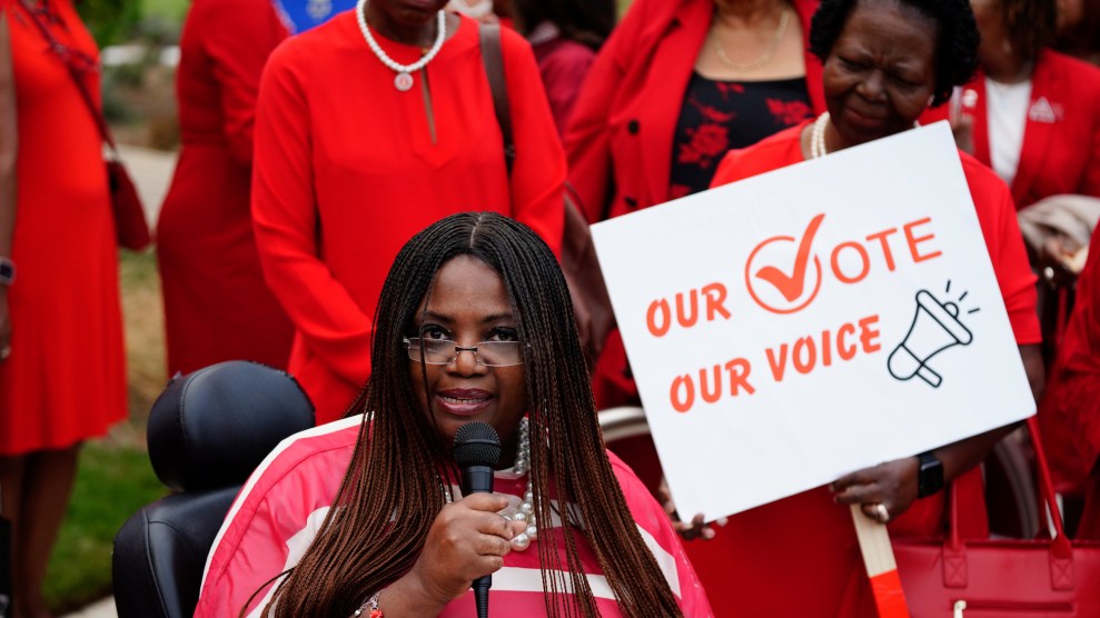 A Black woman wearing red in a wheelchair speaking into a microphone wearing red, surrounded by other Black woman wearing red, including one carrying a poster that says "Our Vote, Our Voice"