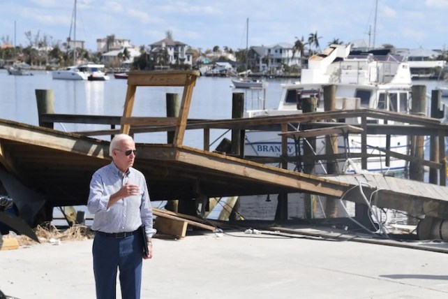 Joe Biden wearing a button down shirt and dress pants speaking with a broken dock behind him