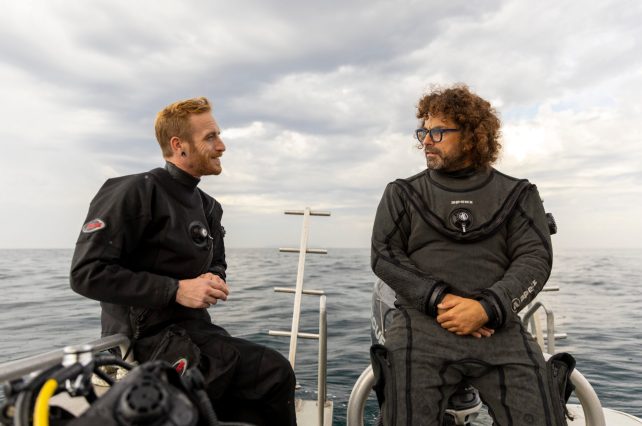 Two individuals sit next to each other on a boat. The one on the right has short red hair and a close cropped beard. The other has longer curly brown hair and a longer beard. Both wear black wetsuits.