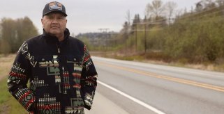 A Native American man in a dark sweater with tribal markings stands by the side of a highway in Washington state.