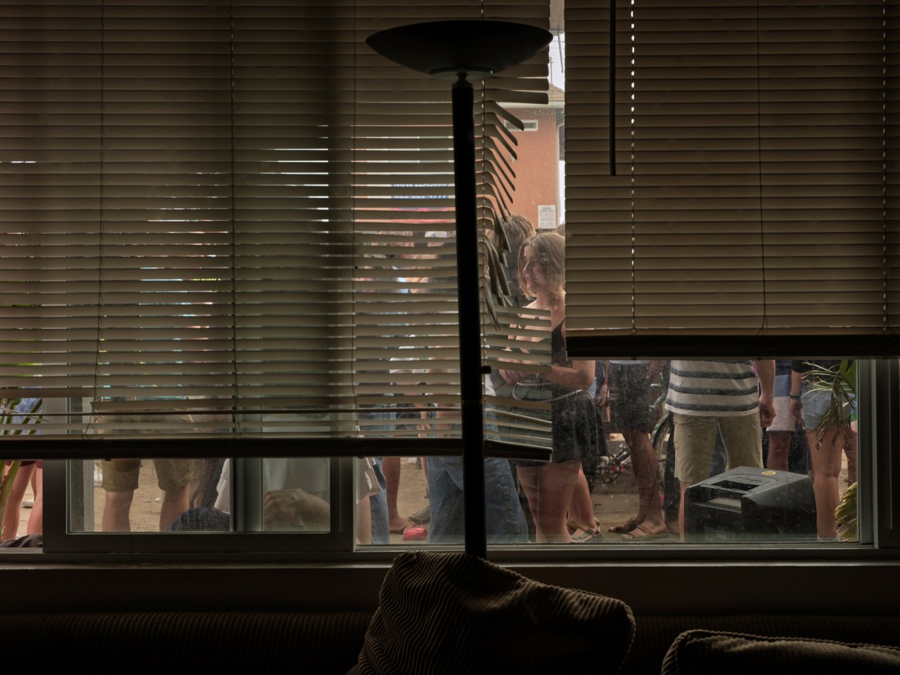 View of a party outside from inside an apartment with blinds partially drawn.
