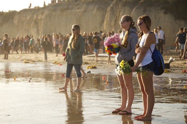 Large group of people standing on the beach looking out at the ocean. Two girls in front holding flowers.