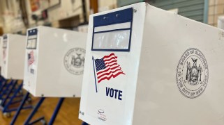 White voting booths that say, "Vote" and "Board of Elections in the City of New York," with an American flag on them, in a school gym