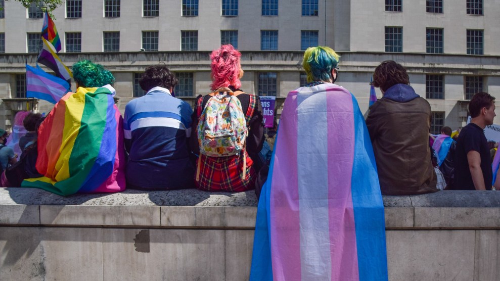 Five individuals sit on a stone bench looking towards a building. You can see their backs, their colorful hair, and the pride flags draped on their soulders.
