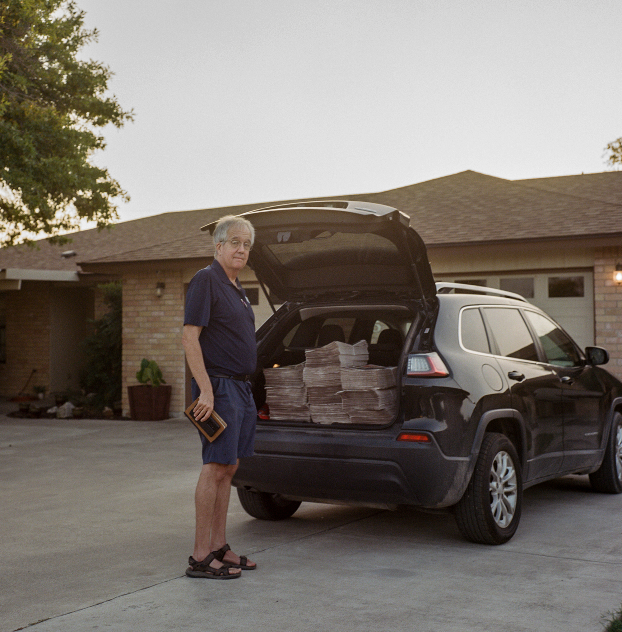 Man standing behind a car with the truck open, newspapers in the truck.