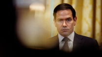 A photo of Marco Rubio dressed in suit and tie staring blankly into the distance toward the camera. He has a sullen expression on his face, and there are yellow-patterned curtains behind him.
