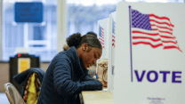 A young African American woman wearing a dark blue coat sits in a folding chair as she fills out her ballot on a table. Erected on the table are several white partitions that have a billowing American flag design on them with the word “Vote” underneath.