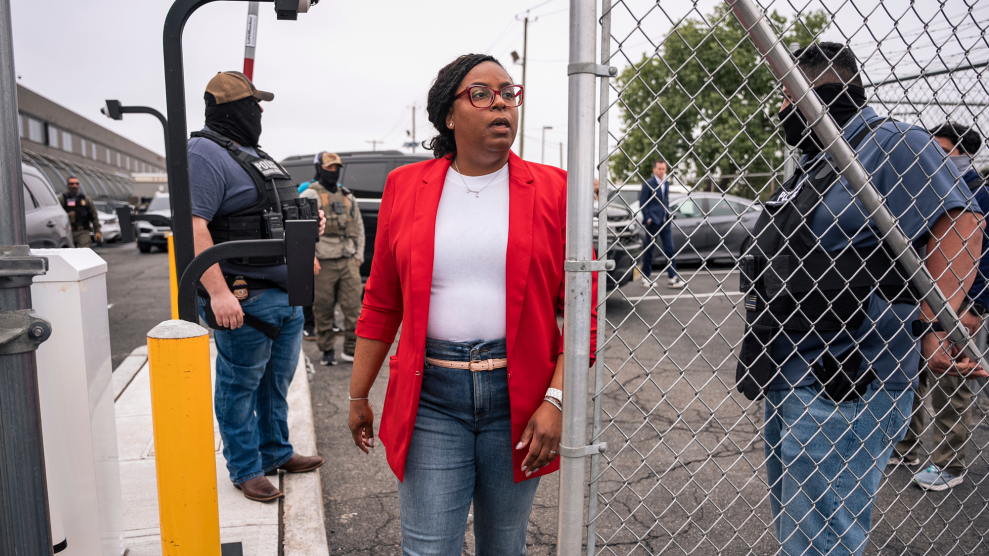 An African American woman, red-rimmed glasses, hair pulled back in braids, walks toward an opening in a chainlink fence. The woman is dressed in a red blazer over a white shirt, and blue jeans. Behind her are agents wearing black vests and face coverings.