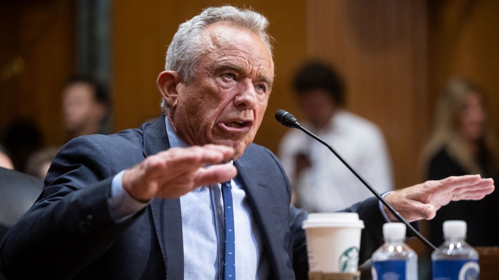 Robert F. Kennedy sits at a microphone with both hands in the air, palms down, as he talks