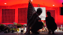 A man kneels while holding the American flag over his right shoulder amid flowers and candles in front of a black-and-white photo of Charlie Kirk. A woman kneels beside him.