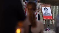 In the foreground, out of focus, is the back of a woman with long hair looking toward a memorial for Charlie Kirk. Beside the memorial, which features a large black-and-white photo of Kirk, stands a man holding an American flag.