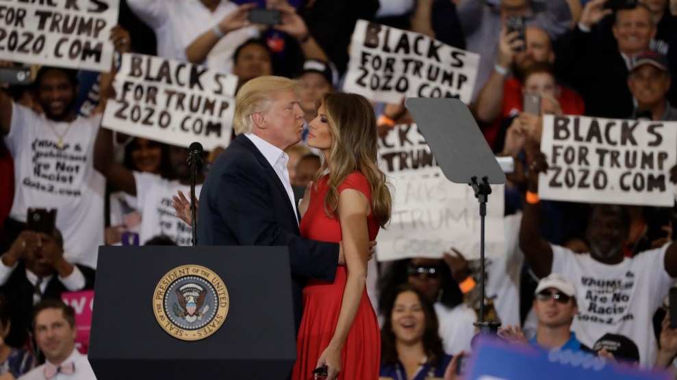 Donald Trump and Melania Trump share a moment on stage at a rally, surrounded by supporters holding 'Blacks for Trump 2020' signs.