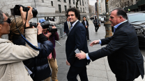 A clean-shaven man with dark curly hair, wearing a dark suit and tie, looks into the camera as photographers and reporters surround him on a New York City sidewalk. A man in a suit behind him holds up his hands, as if attempting to clear a path.