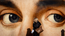 A Caucasian woman with hair down to her shoulders, wearing a coat and carrying two bags, walks to our left as she talks on a mobile phone. Behind her is a large mural of a face, seen from the bridge of the nose to the forehead. The dark gray eyes of the painting seem to be in line with the passing woman, whose shadow falls against the lower part of the wall.