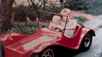 A photo of a toddler and her older brother seated next to one another in a red car on a track that’s laid through a thicket of flowers and bushes. The car, designed for children, is based on an early 20th century racing car.