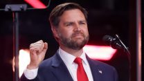 JD Vance wearing a suit with a red tie, holding his fist up on a stage.