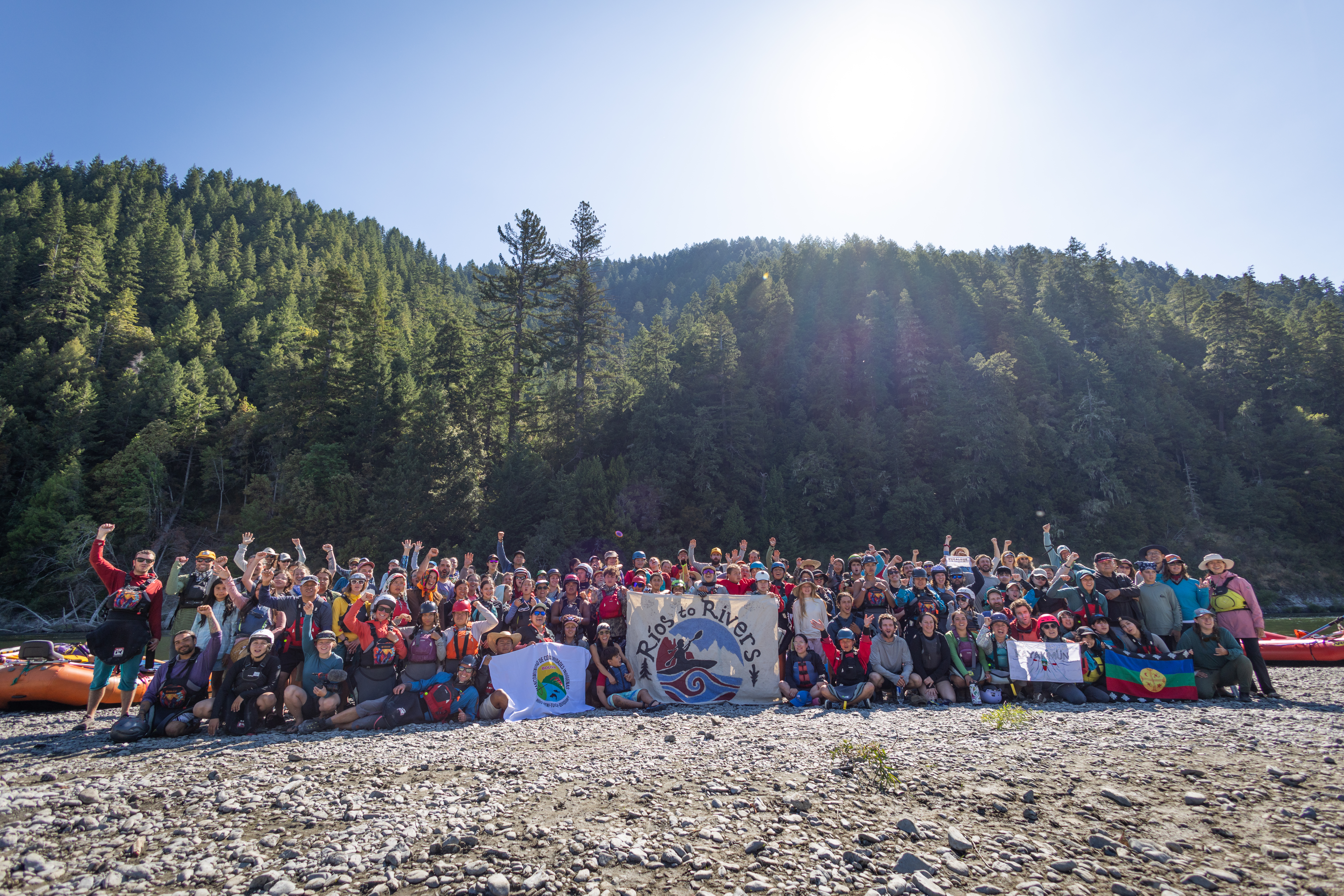 A group of people pose in front of a sign that says Rios to Rivers. Many hold up their fists. They wear gear for kayaking and are standing in front of a river.