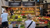 Two people stand in front of a market stall of vegetables.