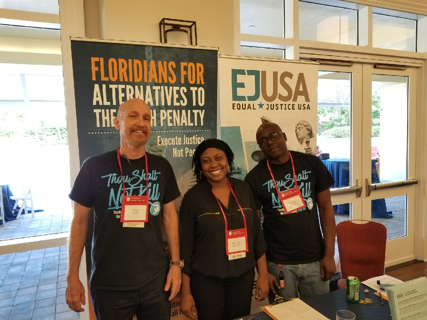 Three EJUSA staff members stand smiling in front of a booth and signage educating people on their organization's mission and the Florida death penalty.