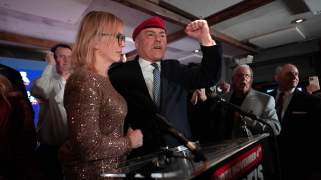 A Caucasian man in a suit and red beret pumps his fist as he stands at a lecturn with supporters gathered around him. Most of the supporters are men in suits. His wife has shoulder-length brown hair and stands beside him in a bronze, glittery dress.
