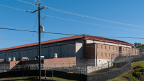 A large rectangular building is surrounded by a tall chain-link fence topped with multiple layers of razor wire. The building is made of brick and has small, narrow windows.
