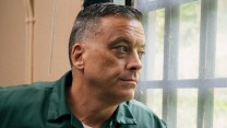 A middle-aged man with a short graying haircut is shown in profile, looking out a barred window, elbow resting on the sill. He wears a green prison jumpsuit, which includes a white tag displaying his name and prisoner identification number.