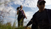 A man in a baseball cap and sunglasses is seen in the foreground as he wades through tall grass. Behind him, in focus, is a young woman wearing glasses, standing on a platform in the grass. She holds a shotgun microphone that’s covered in a fuzzy windscreen and has layers of other equipment strapped around her, including headphones.