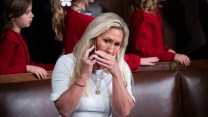 A woman with long blonde hair wearing a white dress, Marjorie Taylor Greene, sits in what appears to be a formal chamber. She is holding a phone to her ear with one hand and covering her mouth with the other, looking concerned or focused. Several children dressed in red clothing stand in the background.