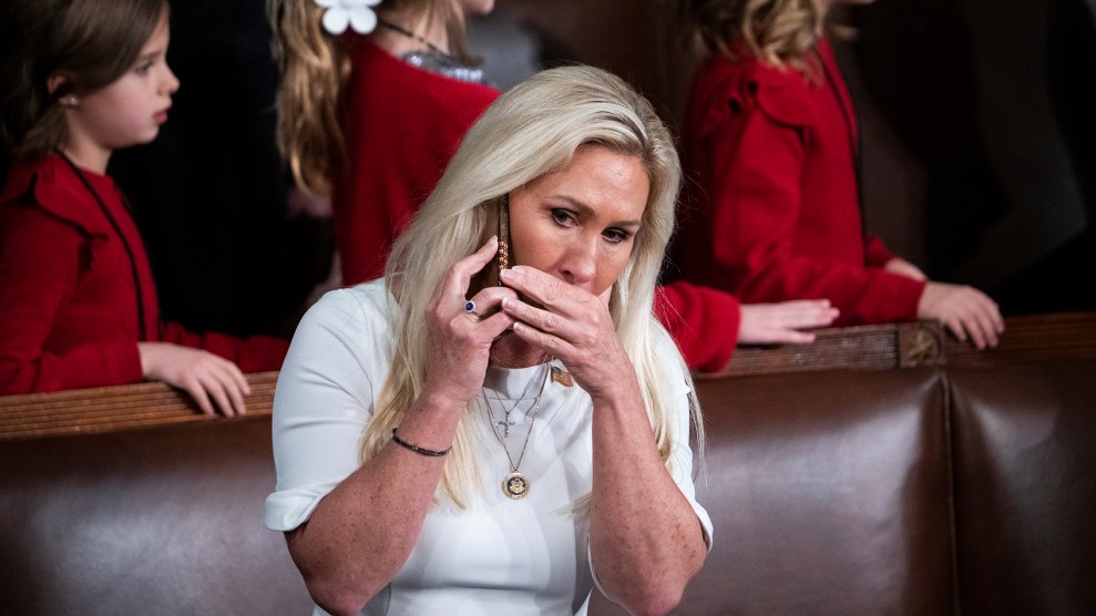 A woman with long blonde hair wearing a white dress, Marjorie Taylor Greene, sits in what appears to be a formal chamber. She is holding a phone to her ear with one hand and covering her mouth with the other, looking concerned or focused. Several children dressed in red clothing stand in the background.