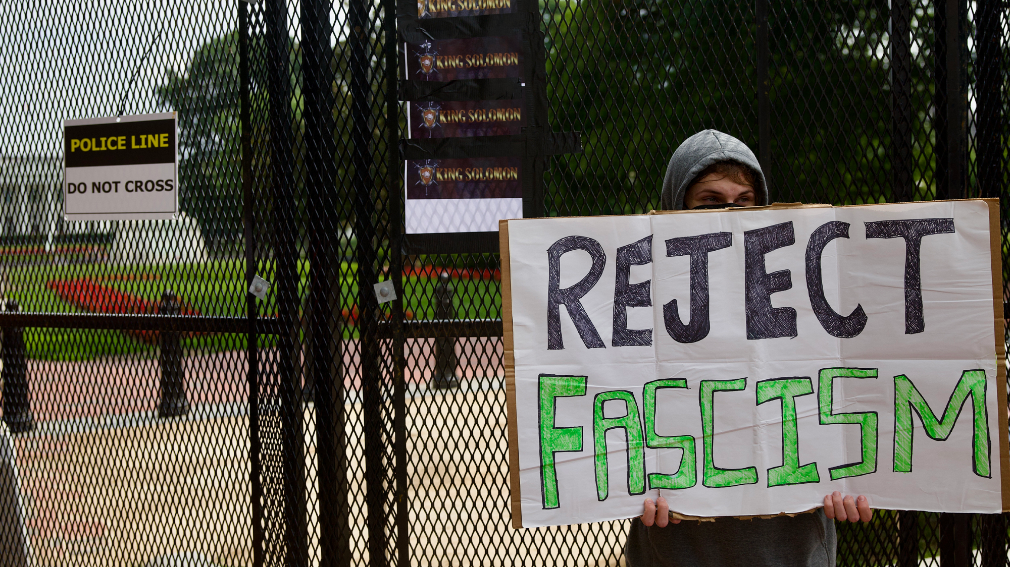 A demonstrator holds a white sign that reads "Reject Facism" in black and green outside the White House. Behind the person is a tall black fence with a sign that reads "Police Line: Do Not Cross."