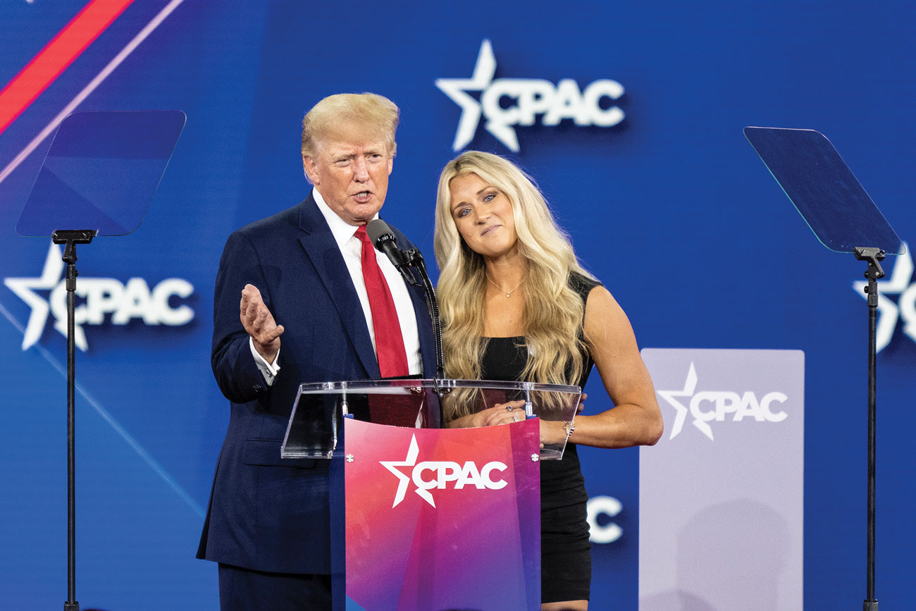 Donald Trump stands on a podium beside a young blond woman wearing a tight black minidress, with a CPAC logo displayed on the wall behind them.