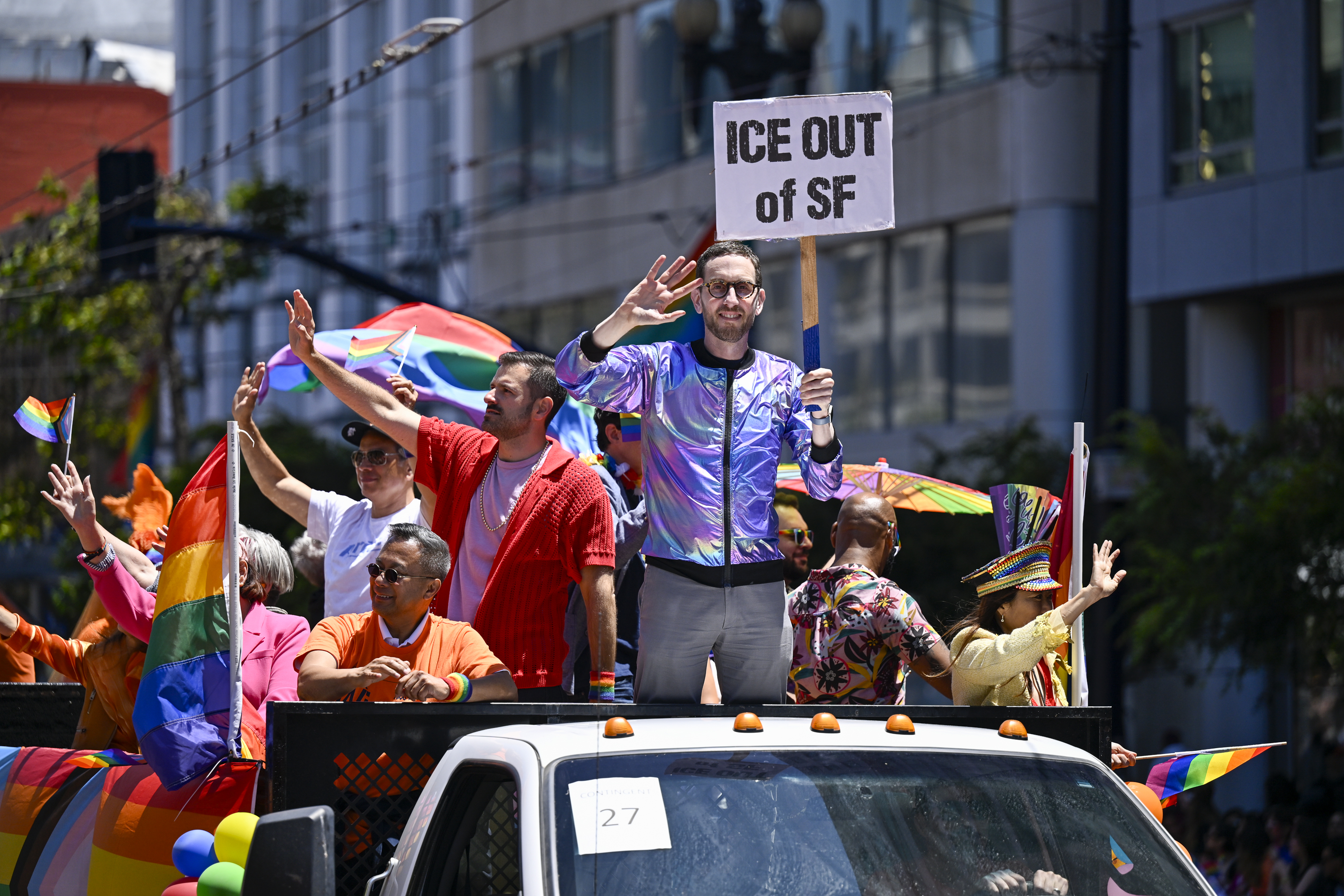 A photo of a man in a colorful jacket waving with one hand as he holds a sign in the other. The sign reads, "ICE OUT of SF." The man, who is wearing glasses and who has a light beard, stands on the back of a truck with flags alongside other people who are dressed in bright colors and are also waving to the crowd.