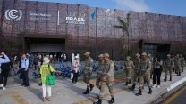 A group of people in grey-green uniforms walk past a building with a sign that says "COP30 BRASIL AMAZONIA"