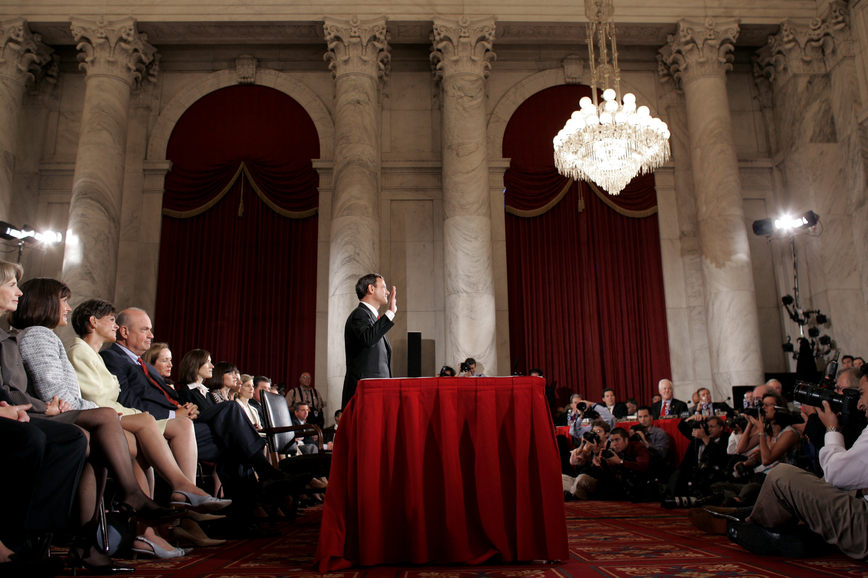 John Roberts raises his right hand in a large hearing room as he faces a crowd of photographers and members of the Senate Judiciary Committee.