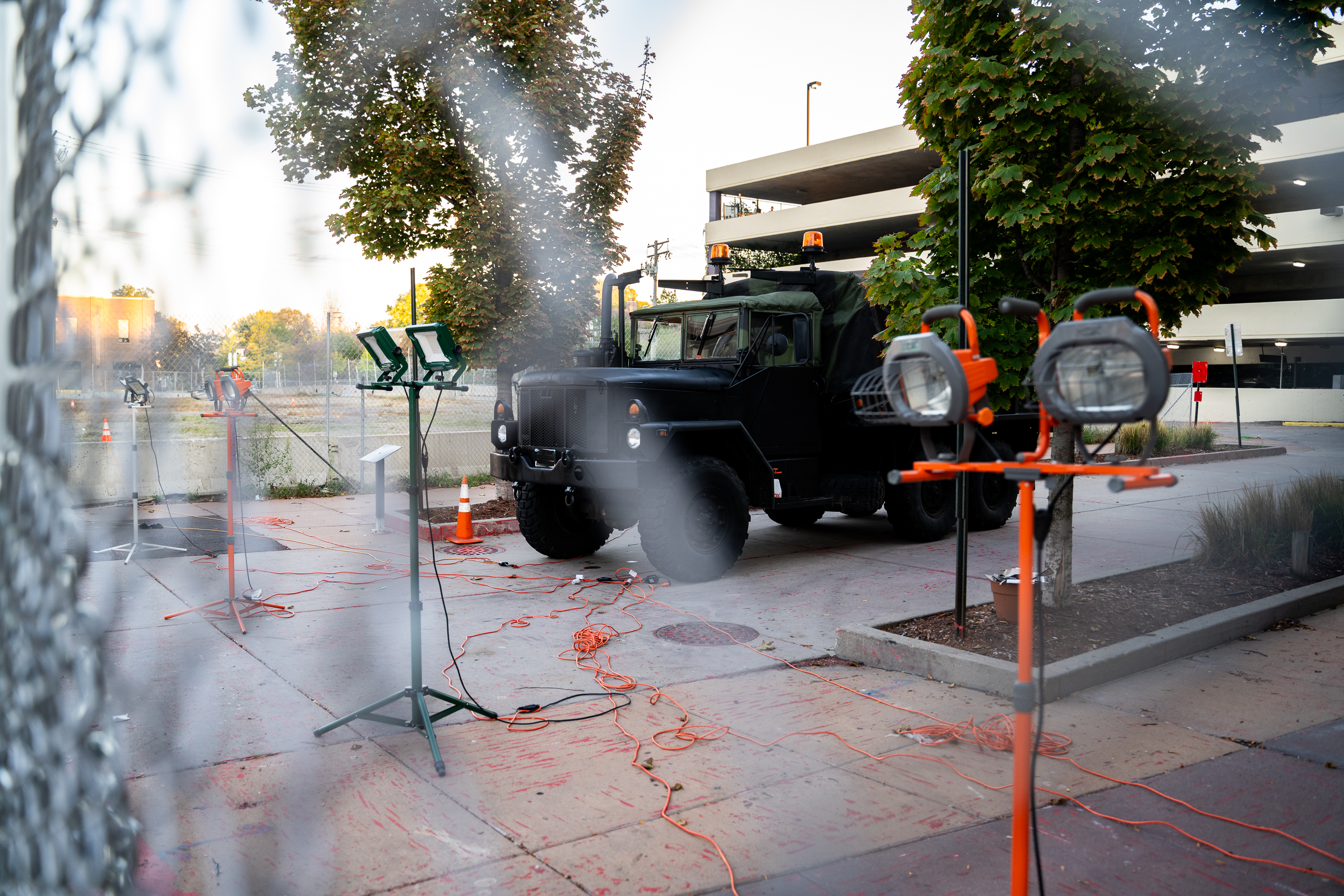 A military style truck and floodlights positioned behind a chainlink fence near a mall parking garage.