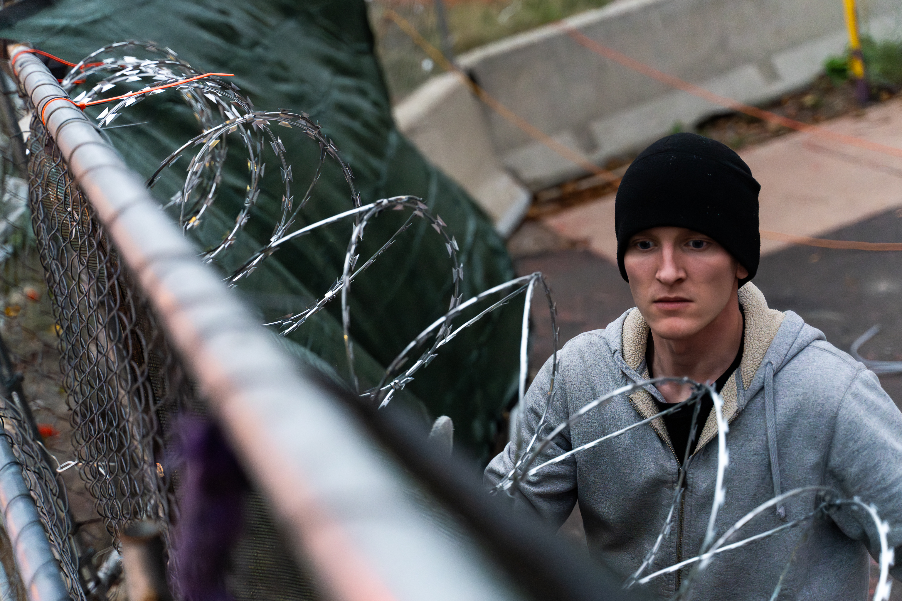 A young man in a sweatshirt and knit cap surveys his work installing razor wire.