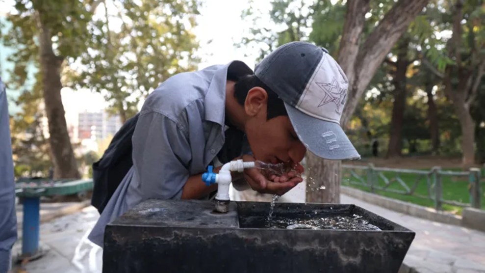 A young person wearing a baseball cap crouches over a water fountain drinking water.