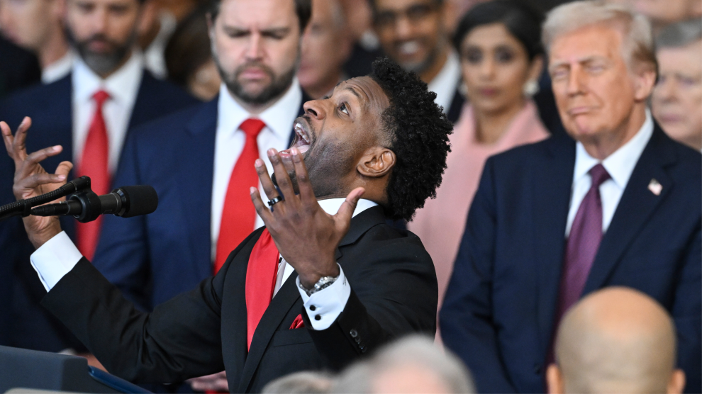 A middle-aged Black man in a dark suit and red tie stands behind a presidential lectern, speaking with hands raised as he looks upward. Behind him is a crowd mostly of men in suits, among them Donald Trump and JD Vance, who stand with heads slightly bowed and eyes closed.