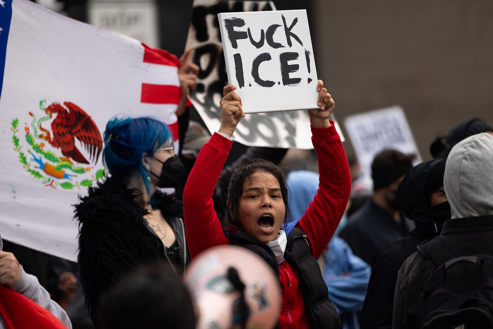 A woman in a crowd, who appears to be shouting, holds a sign that reads, "Fuck ICE."