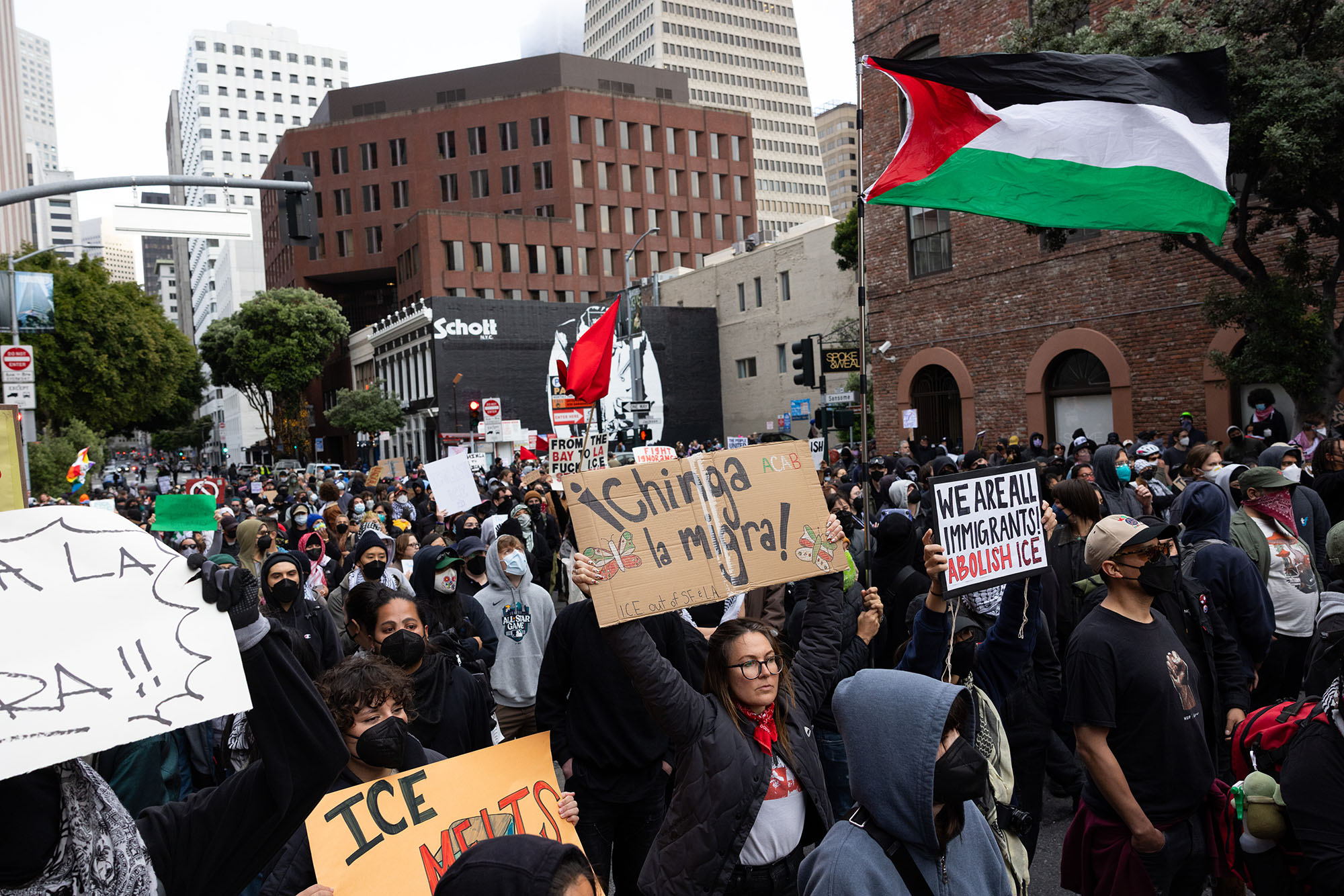 A large crowd marches down a street in San Francisco. In the foreground, two people carry signs that read, "Chinga la migra!" and "We are all immigrants! Abolish ICE."