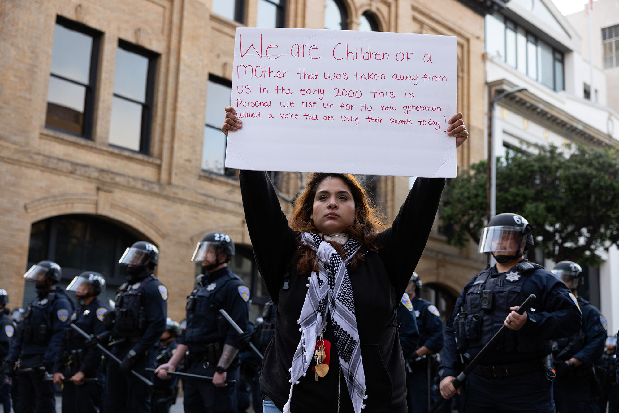 A woman stands in front of a line of police in riot gear, holding a handwritten sign above her head. The sign reads: "We are children of a mother that was taken away from us in the early 2000 [sic]. This is personal. We rise up for the new generation without a voice that are losing their parents today!"