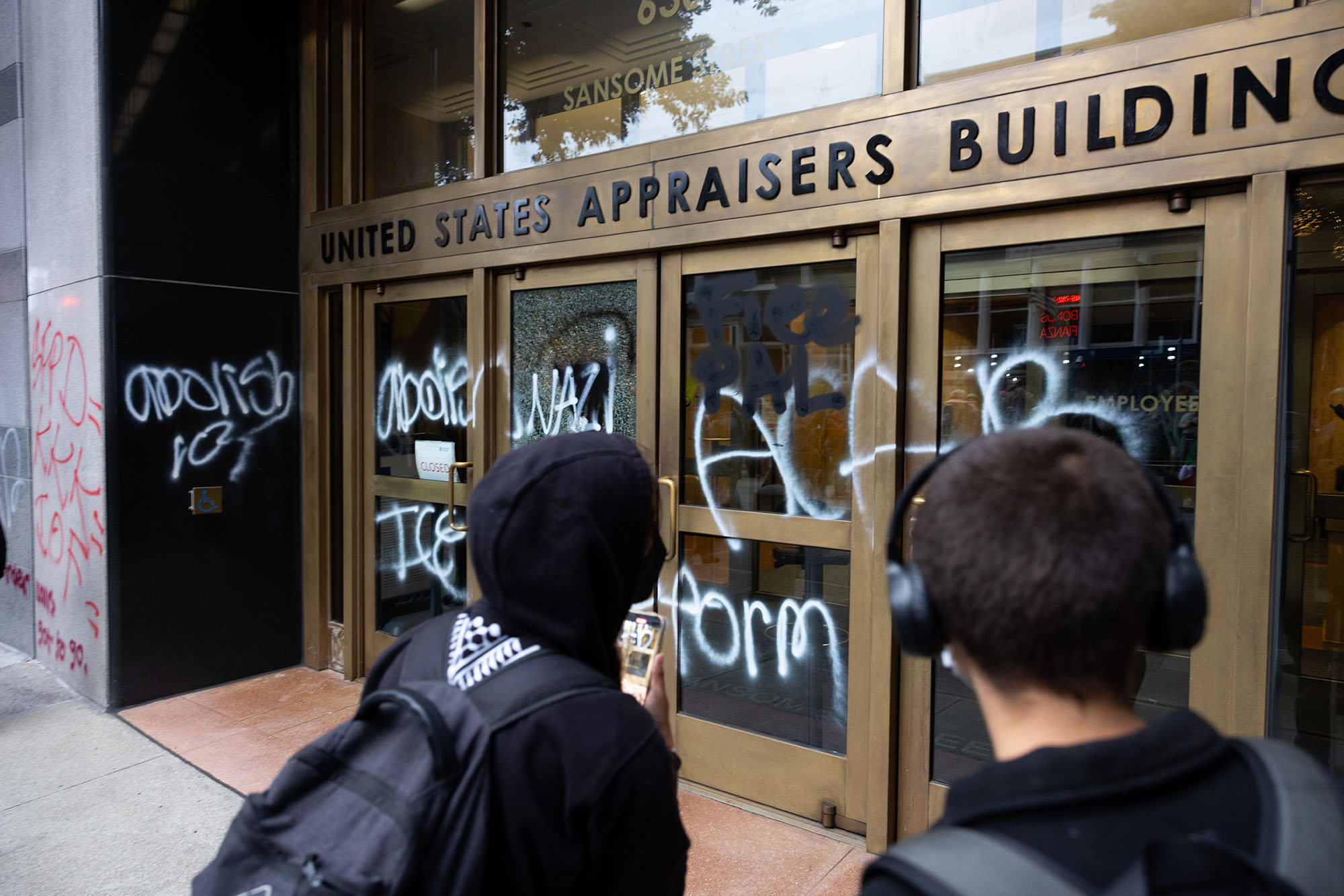 Two people, seen from behind, look at graffiti spray-painted on the stone facade and glass doors of a building.