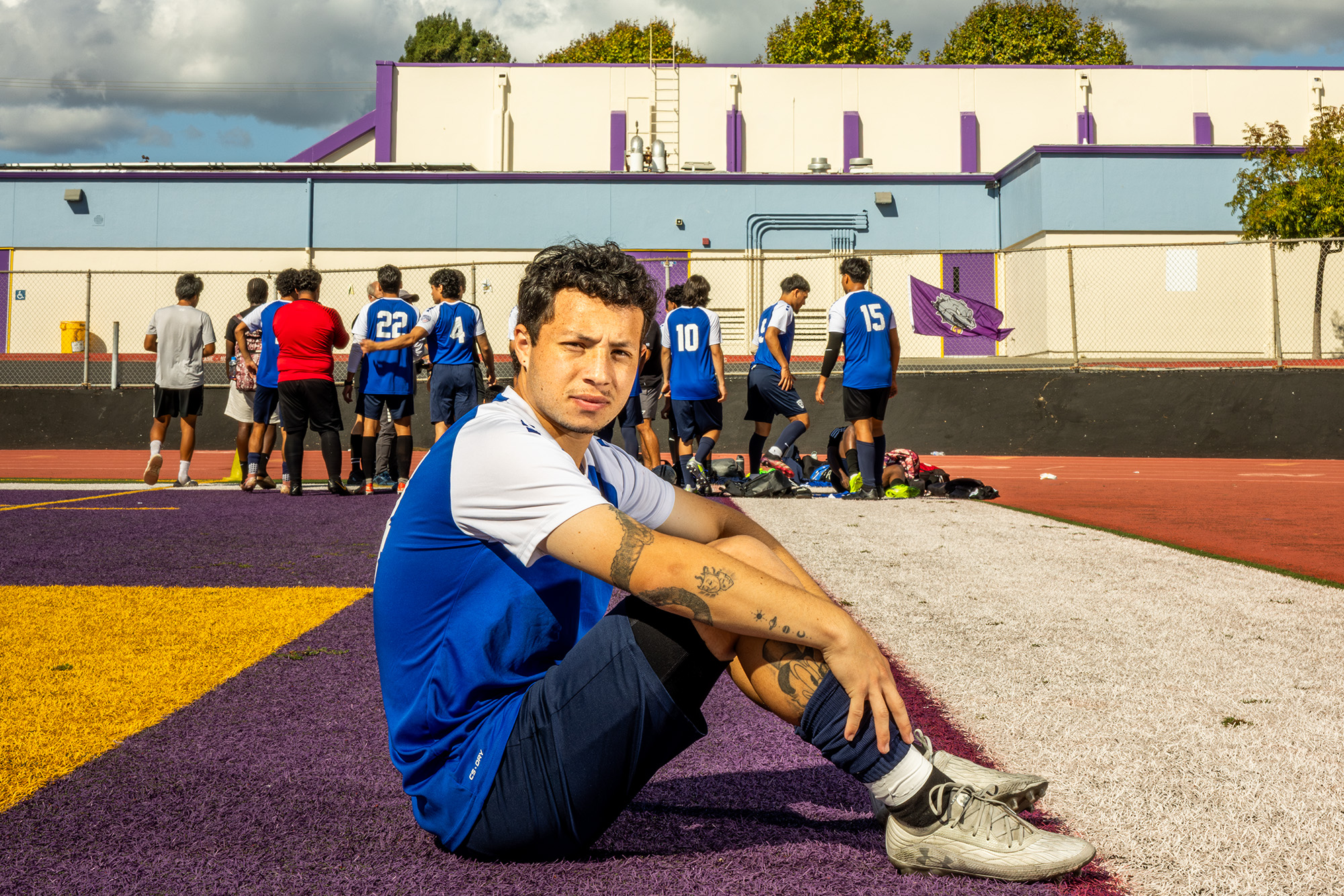 Dilan Pinzón sedí na astroturfovaném fotbalovém hřišti. Má na sobě modrý dres a bílé kopačky a zírá přímo do kamery. Ostatní hráči stojí v pozadí zády ke kameře.