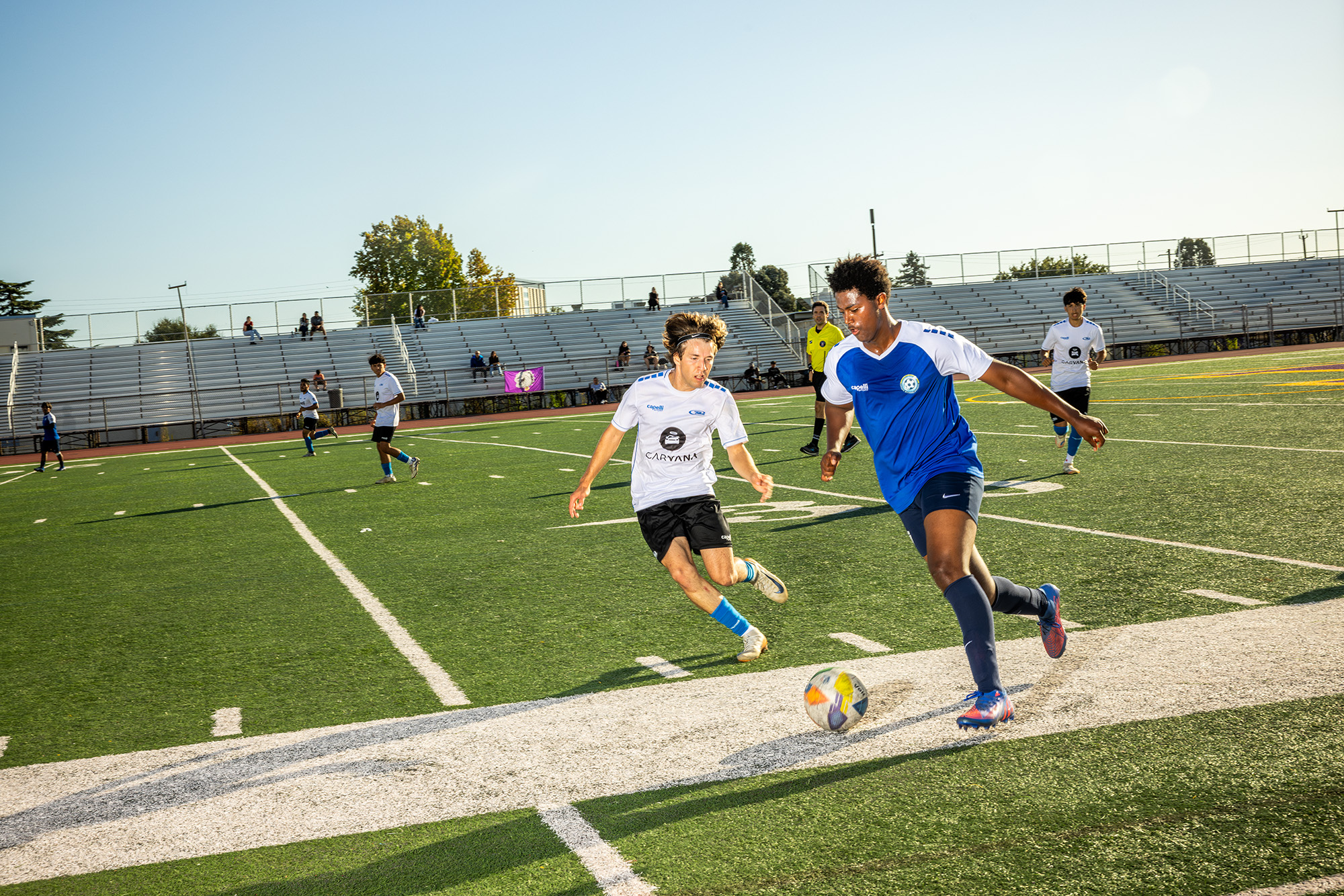 Dva fotbalisté – jeden v bílém dresu a druhý v modrém – kopnou do fotbalového míče na zeleném astroturfovaném hřišti.